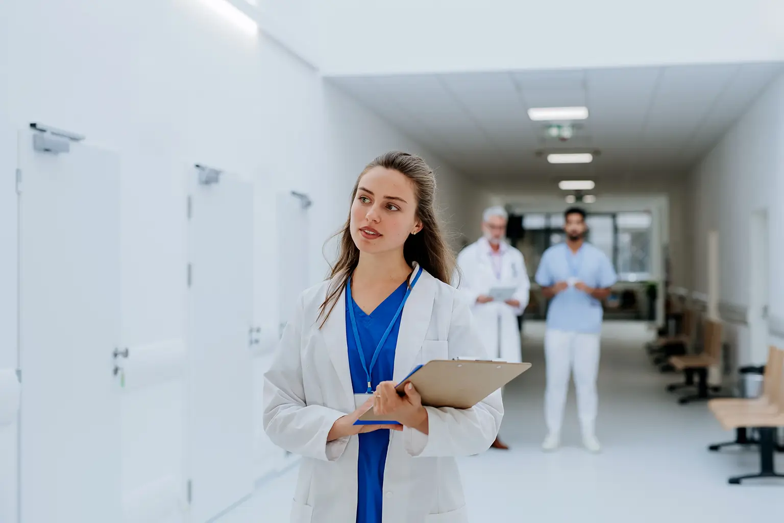 A female doctor stands in a busy hallway with other people, indicating a collaborative healthcare environment.